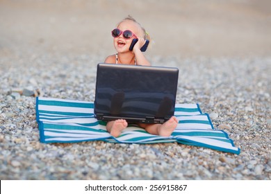 A cute little happy girl in pink sunglasses sitting on the striped blue and white mat with a laptop on the beach on a warm summer day. Holiday on a seashore concept. Funny kids. Sea. Ocean.