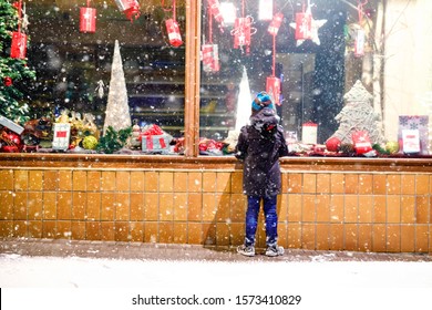 Cute Little Healthy School Kid Boy On Christmas Market. Funny Happy Child In Fashion Winter Clothes Making Window Shopping Decorated With Gifts, Xmas Tree. Snow Falling Down, Snowfall