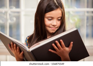 Cute Little Hispanic Girl Reading A Story From A Big Book In The Living Room