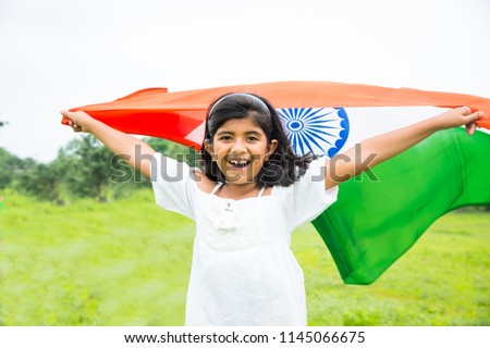 cute little Indian girl holding, waving or running with Tricolour near a lake with greenery in the background, celebrating Independence or Republic day  

