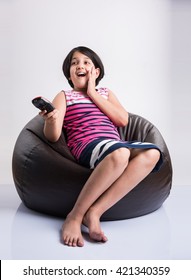 Cute Little Indian Girl Watching Television / TV While Changing Channels Using Remote Control, Sitting Over Bean Bag, Isolated Over White Background