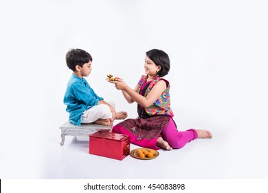 Cute little Indian Sister tying Rakhi to Her little brother's wrist and exchanging gifts and sweets on Raksha Bandhan or Bhai Dooj festival, Sitting isolated over white background