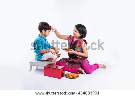 Cute little Indian Sister tying Rakhi to Her little brother's wrist and exchanging gifts and sweets on Raksha Bandhan or Bhai Dooj festival, Sitting isolated over white background