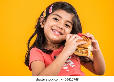 Cute Little Indian/Asian Girl Child Eating Tasty Burger, Sandwich Or Pizza In A Plate Or Box. Standing Isolated Over Blue Or Yellow Background.