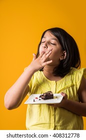 Cute Little Indian/Asian Girl Child Eating Piece Of Strawberry Or Chocolate Flavoured Pastry/cake In A Plate Or Licking Fingers. Isolated Over Colourful Background