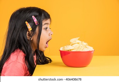 Cute Little Indian/asian Girl Kid Eating Chips Or Potato Wafers In Big Red Bowl, Over Yellow Background