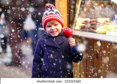 Cute Little Kid Boy Eating Sweet Red Apple On German Christmas Market. Happy Child In Winter Clothes With Lights On Background. Kid Looking At The Camera. Family, Tradition, Holiday Concept