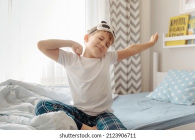 Cute Little Kid Boy Wake Up In The Morning In His Bed, Stretching Hand Rise Up To The Air While Sitting In Sunny Bedroom With Big Window On Background.