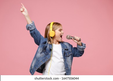 Cute little kid girl 12-13 years old isolated on pastel pink background. Childhood lifestyle concept. Mock up copy space. Listen music with headphones sing song in microphone pointing index finger up
