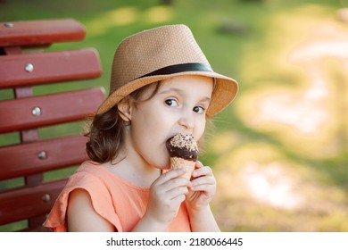 Cute Little Kid Girl In A Hat Eating Ice Cream Cone. Child Sitting On A Park Bench And Laughing . Summer Concept.