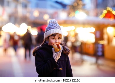 Cute Little Kid Girl Having Fun On Traditional Christmas Market During Strong Snowfall. Happy Child Eating Traditional Curry Sausage Called Wurst. Schoolgirl Standing By Illuminated Xmas Tree.