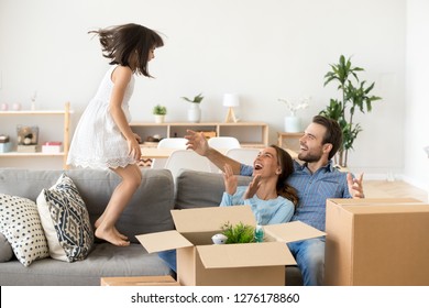Cute little kid girl jumping on sofa excited on moving day, family mom dad child laughing having fun in new home unpacking boxes, happy daughter playing helping parents to pack enjoying relocation
