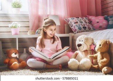 Cute Little Kid Girl Is Reading A Book At Home. Funny Lovely Child Having Fun In Kids Room. 