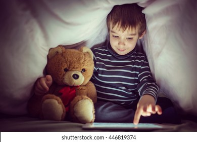 Cute little kid with his friend teddy bear using tablet computer under blanket at night in a dark room