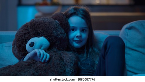 Cute Little Kid Sitting On Couch With Teddy Bear While Watching Tv At Home In Evening. Portrait Of Preschool Girl Hugging Stuffed Animal Sitting On Sofa And Watching Cartoon Late At Night