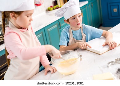 Cute Little Kids In Chef Hats Making Dough And Reading Cookbook In Kitchen