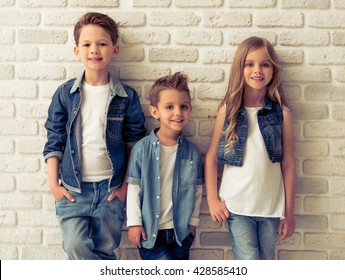 Cute little kids in stylish jeans clothes are looking at camera and smiling, standing against white brick wall