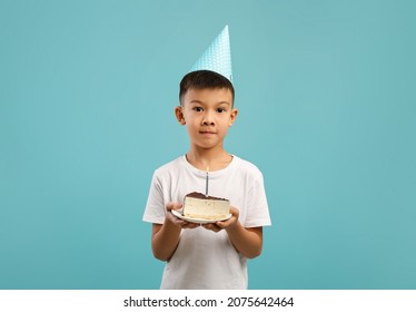 Cute Little Korean Boy Wearing Party Hat Holding Piece Of Birthday Cake With Candle And Looking At Camera, Happy Asian Male Child Celebrating His B-Day, Posing Over Blue Studio Background, Copy Space