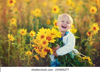 A Cute Little Laughing Girl In The Field Of Sunflowers Holding A Huge Bunch Of Flowers In A Sunny Summer Evening. 