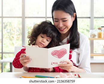 Cute Little Mixed Race Girl Sitting On Beautiful Asian Mother's Lap Looking At The Card Written LOVE MOM In The Kitchen. 