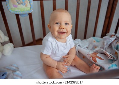 A cute little newborn baby is sitting in a crib, drinking water from a bottle. a little boy is sitting in a crib and smiling. The concept of childhood