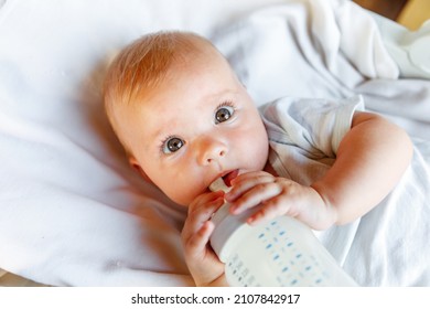 Cute Little Newborn Girl Drinking Milk From Bottle And Looking At Camera On White Background. Infant Baby Sucking Eating Milk Nutrition Lying Down On Crib Bed At Home. Motherhood Happy Child Concept