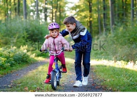 Cute little preschool girl in safety helmet riding bicycle. School kid boy, brother teaching happy healthy sister child cycling and having fun with learning bike. Active siblings family outdoors.