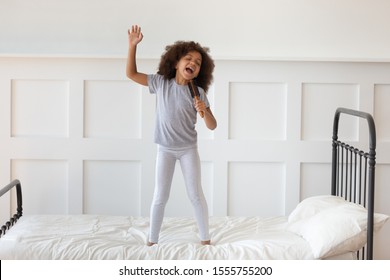 Cute Little Preschooler African American Girl Stand On Bed Sing In Toy Hairbrush Microphone, Funny Small Artistic Biracial Kid Have Fun Playing At Home In Morning, Perform Entertain In Bedroom