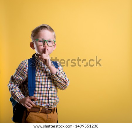 Cute little school boy asking and make sign to keep silence. Child isolated on yellow background. Secrets of success knowledge and education. Innovation in primary school.