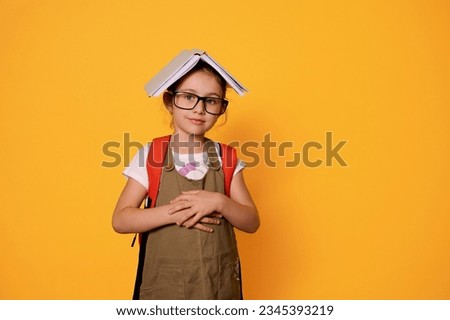 Cute little school kid, child girl with a book on her head like a roof, looks confidently at camera, carrying a backpack, isolated over orange studio background. People. Erudition. Education Knowledge