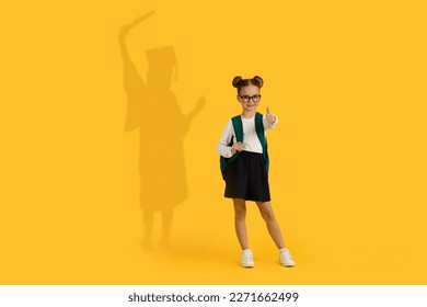 Cute Little Schoolgirl With Backpack Showing Thumb Up, Having Shadow In Graduation Costume On Her Back Over Yellow Background, Smiling Female Pupil Child Dreaming About High Education, Collage - Powered by Shutterstock