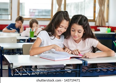 Cute little schoolgirls studying together at desk with classmates in background