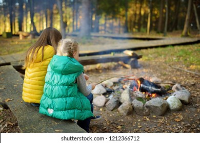 Cute Little Sisters Roasting Hotdogs On Sticks At Bonfire. Children Having Fun At Camp Fire. Camping With Kids In Fall Forest. Family Leisure With Kids At Autumn.