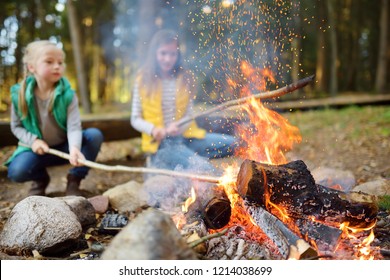 Cute Little Sisters Roasting Hotdogs On Sticks At Bonfire. Children Having Fun At Camp Fire. Camping With Kids In Fall Forest. Family Leisure With Kids At Autumn.