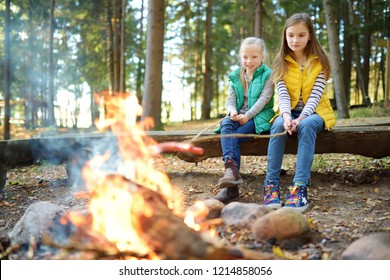 Cute Little Sisters Roasting Hotdogs On Sticks At Bonfire. Children Having Fun At Camp Fire. Camping With Kids In Fall Forest. Family Leisure With Kids At Autumn.