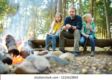 Cute Little Sisters And Their Father Roasting Marshmallows On Sticks At Bonfire. Children Having Fun At Camp Fire. Camping With Kids In Fall Forest. Family Leisure With Kids At Autumn.