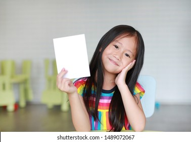 Cute little smiling Asian child girl holding blank white paper card in her hand. Kid showing empty paper note copy space in children room.