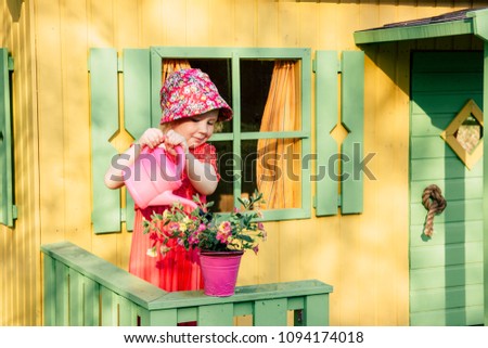 Cute little smiling gardener girl wearing floral summer hat watering pretty pink flowers with pink watering can in front of a playhouse.