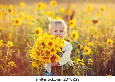 A Cute Little Smiling Girl In The Field Of Sunflowers Holding A Huge Bunch Of Flowers In A Sunny Summer Evening. 