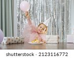 Cute little smiling girl with short curly hair and stars on face in pink poofy dress lift pink balloon near fruit cake.