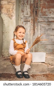 Cute little smiling shy girl sitting on a ladder bench next to old house. Close up portrait of happy three years old child with two blonde pigtails. Rustic, retro, vintage style outfit.