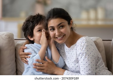 Cute little son telling secret to cheerful Indian mom, whispering in ear. Happy loving mother and kid enjoying being friendship, trust, spending leisure time together, having fun, talking at home