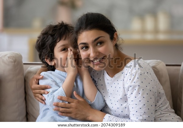 Cute little son telling secret to cheerful Indian\
mom, whispering in ear. Happy loving mother and kid enjoying being\
friendship, trust, spending leisure time together, having fun,\
talking at home