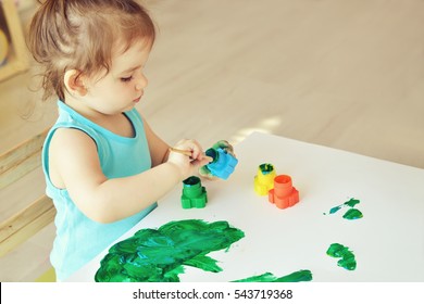 Cute Little Toddler Child Painting With Paintbrush And Colorful Paints. Adorable Baby Girl Drawing On White Paper Near Window In Light Room