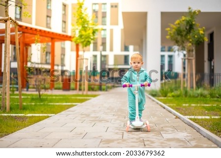 Cute little toddler girl in blue overalls riding on kick scooter. Happy healthy lovely baby child having fun in the city. Active kid on cold day outdoors. High quality photo