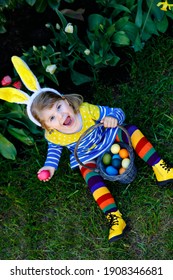 Cute Little Toddler Girl With Bunny Ears Having Fun With Traditional Easter Eggs Hunt On Warm Sunny Day, Outdoors. Happy Child Celebrating Family Christian Holiday With Basket With Colored Egg