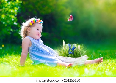 Cute little toddler girl with curly hair wearing a blue summer dress having fun watching a butterfly and flowers, relaxing in the garden on a sunny spring day
