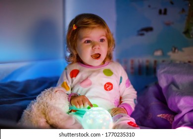 Cute Little Toddler Girl Playing With Colorful Night Light Lamp Before Going To Bed. Sleepy Tired Baby Daughter In Nightwear Having Fun. Healthy Child Not Sleeping.