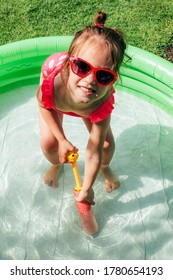 Cute Little Toddler Girl Smiling Playing In Inflatable Water Pool On Sunny Day Outdoor. Summer Leisure Concept. Happy Childhood. Genuine Lifestyle Moments.