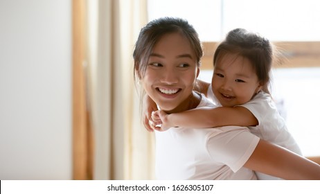 Cute Little Vietnamese Girl Piggyback Overjoyed Ethnic Young Mother Having Fun At Home, Small Smiling Asian Daughter Child Play With Happy Biracial Millennial Mom Or Nanny, Entertainment Concept